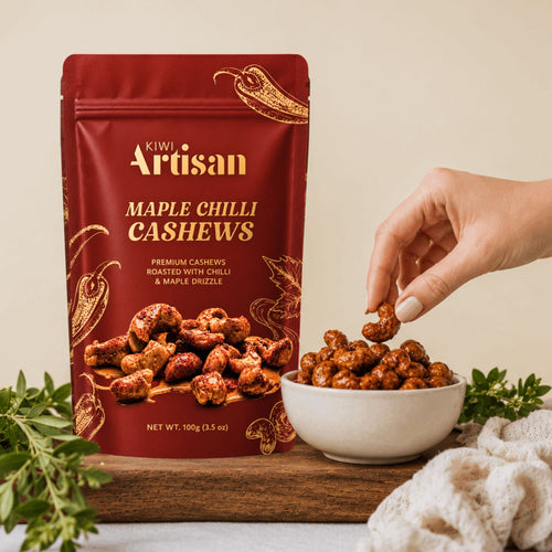 A stand-up pouch of Kiwi Artisan Maple Chilli Cashews stands on a wooden board next to a white bowl filled with the roasted cashews, with a hand reaching in to pick one. Fresh herbs and a cream knit cloth are arranged around the product against a warm beige background.