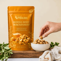 A stand-up pouch of Kiwi Artisan Manuka Honey Macadamias stands on a wooden surface next to a white bowl filled with the nuts, with a hand reaching in to grab one. Fresh green herbs and a soft beige cloth are arranged around the setup against a warm cream background.