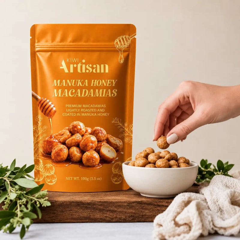 A stand-up pouch of Kiwi Artisan Manuka Honey Macadamias stands on a wooden surface next to a white bowl filled with the nuts, with a hand reaching in to grab one. Fresh green herbs and a soft beige cloth are arranged around the setup against a warm cream background.