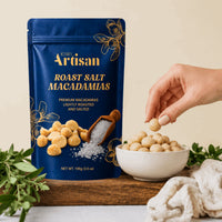 A blue stand-up pouch of Kiwi Artisan Roast Salt Macadamias stands on a wooden board next to a white bowl filled with the macadamias, with a hand reaching in to pick one. Green leaves and a beige cloth are arranged around the setup.