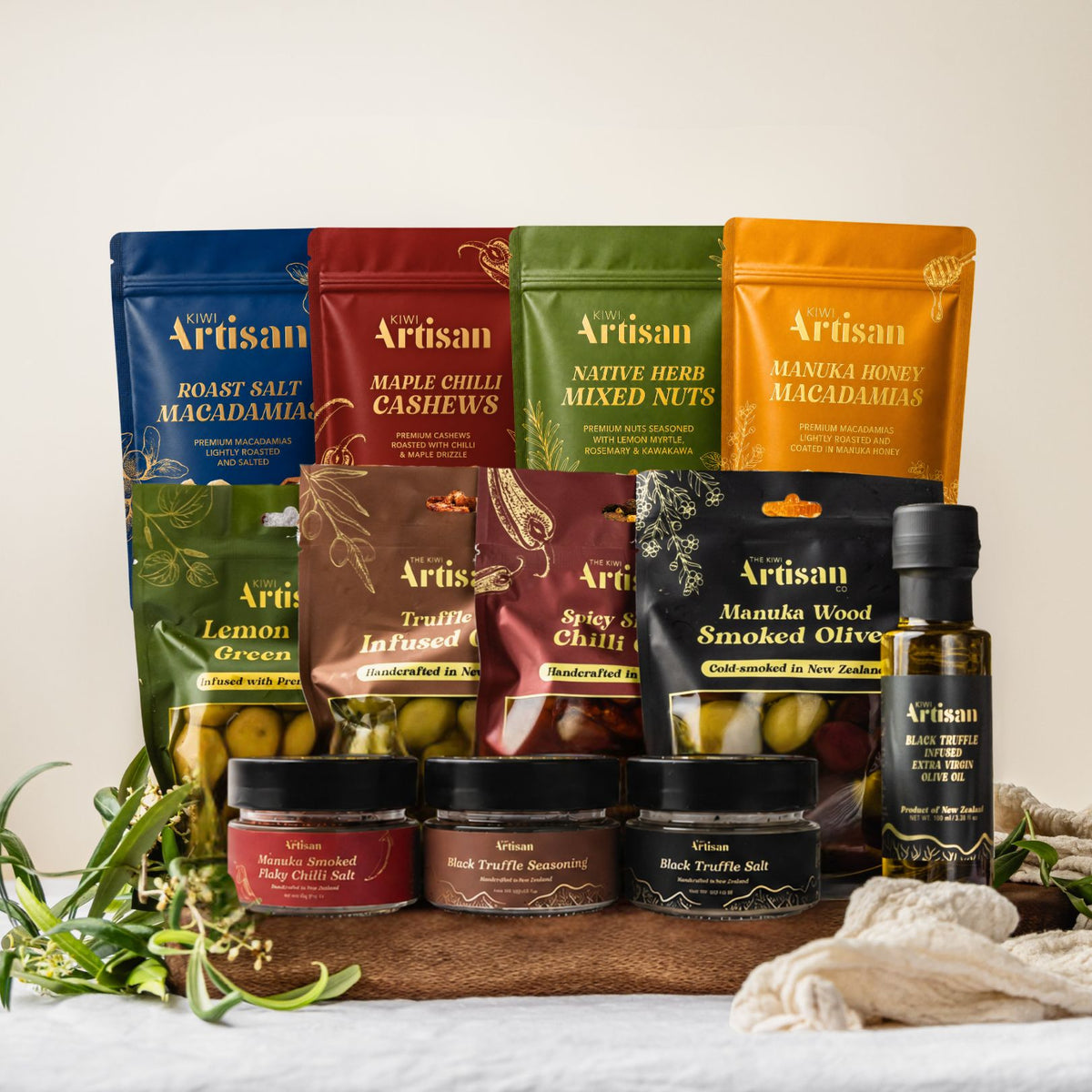 A curated gift set of Artisan food products arranged on a wooden board with decorative herbs and a cloth. The set includes colorful stand-up pouches of roasted salt macadamia, mixed nuts, honey macadamia, lemon green olives, stuffed chilis, smoked olives, jars of chili salt, truffle salt, and a bottle of extra virgin olive oil.