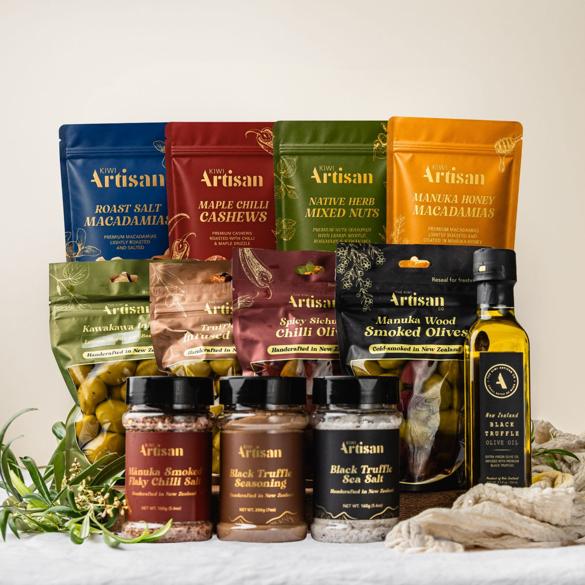 A curated gift assortment of Artisan brand food products, including roasted salt macadamia bags, cashews, mixed nuts, spices, sea salt, and olive oil, arranged on a white surface with decorative herbs and rope.