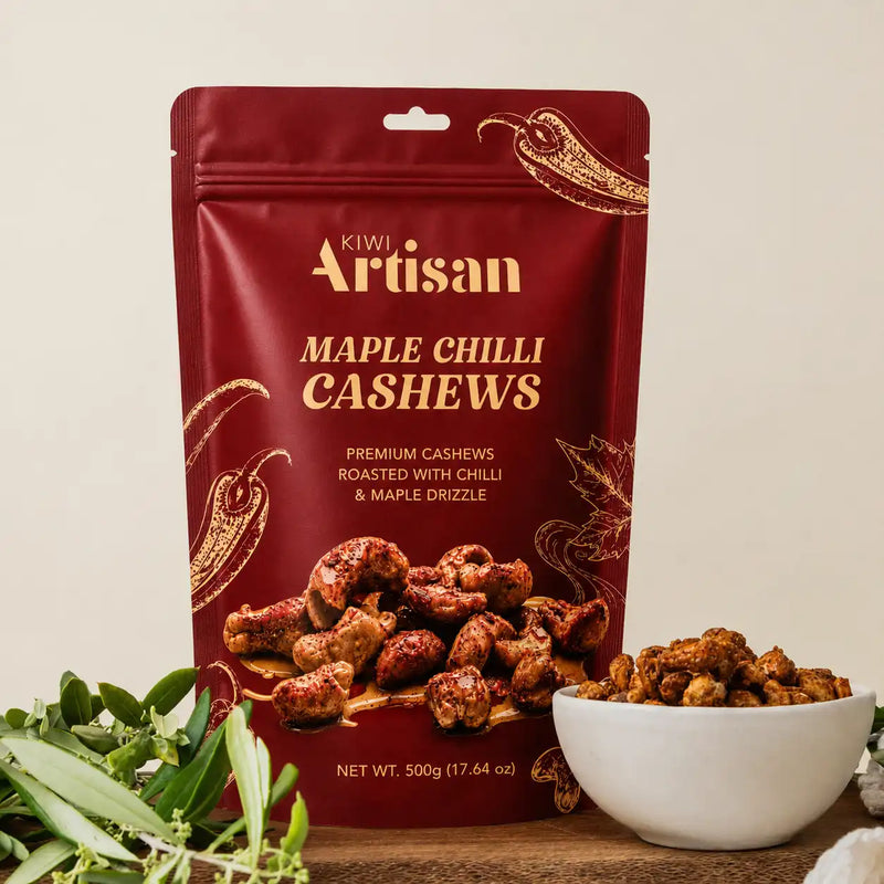 A 500g stand-up pouch of Kiwi Artisan Maple Chilli Cashews stands on a wooden surface with a small white bowl of the roasted cashews and green herbs in front. The pouch is deep red with gold lettering and decorative chili illustrations.