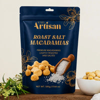 A 500g stand-up pouch of Kiwi Artisan Roast Salt Macadamias, displayed on a wooden surface with a bowl of the raw macadamias and greenery in the background.