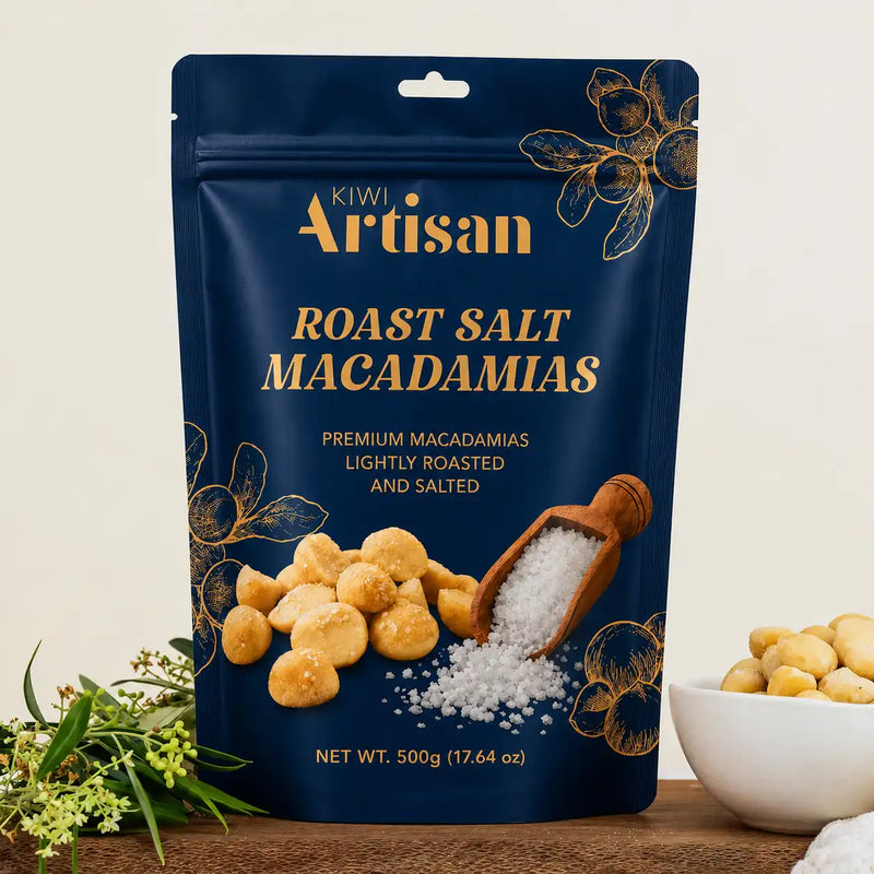 A 500g stand-up pouch of Kiwi Artisan Roast Salt Macadamias, displayed on a wooden surface with a bowl of the raw macadamias and greenery in the background.