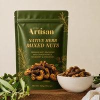A 500g bag of Kiwi Artisan Native Herb Mixed Nuts in a dark green stand-up pouch with gold lettering, displayed next to a white bowl of the mixed nuts on a wooden surface with green leaves.