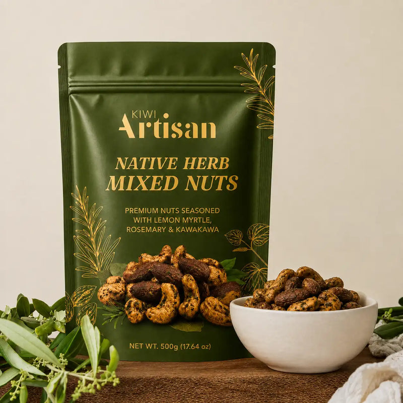 A 500g bag of Kiwi Artisan Native Herb Mixed Nuts in a dark green stand-up pouch with gold lettering, displayed next to a white bowl of the mixed nuts on a wooden surface with green leaves.