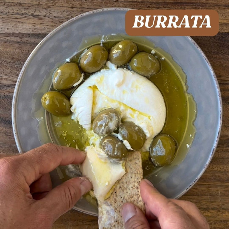 Burrata with olives on a plate, with a hand holding bread with cheese.