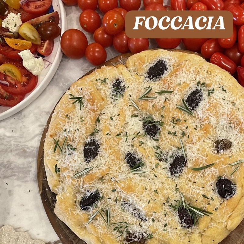 Focaccia bread with olives and herbs on a marble surface with tomatoes and a bowl of salad.